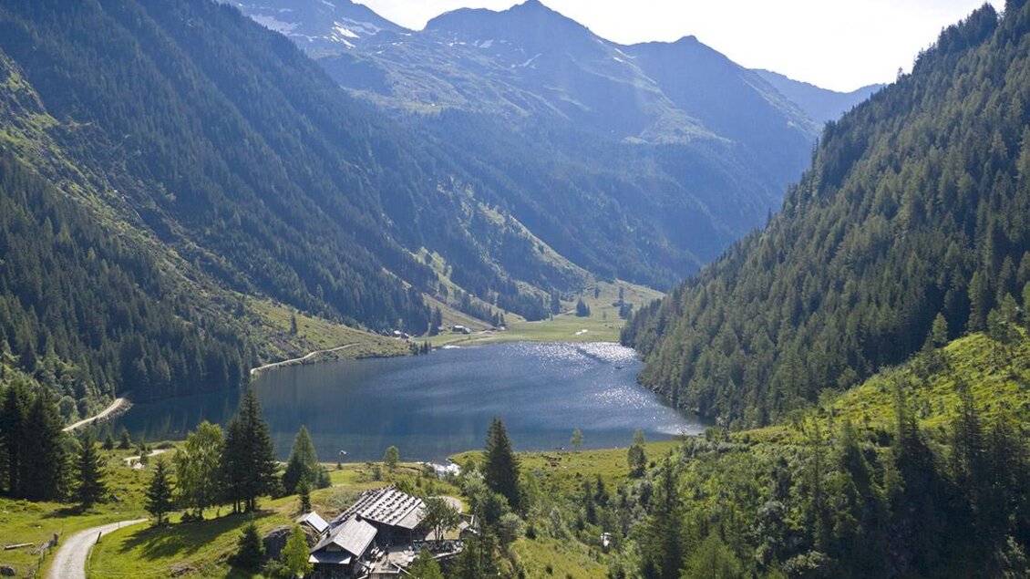 Eine malerische Landschaft mit Bergen und einem klaren See. Im Vordergrund ist ein charmantes Holzhaus zu sehen, umgeben von üppigem Grün. | © Herbert Raffalt