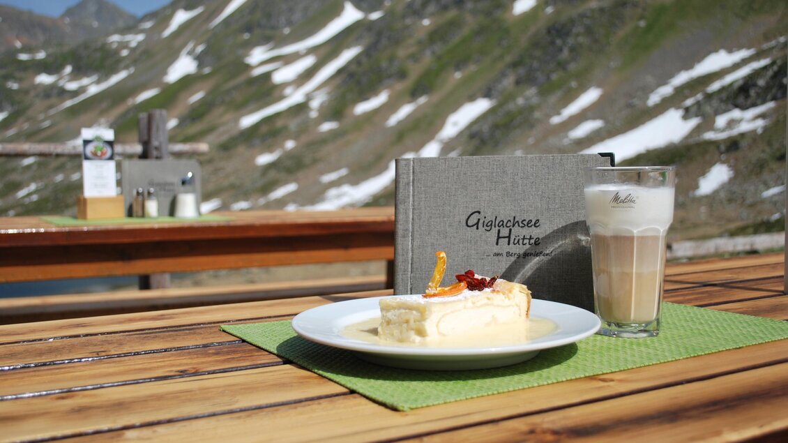 Ein Stück Kuchen mit frischen Früchten und ein Glas Latte Macchiato auf einem Holztisch. Im Hintergrund sind schneebedeckte Berge zu sehen. | © Giglachseehütte