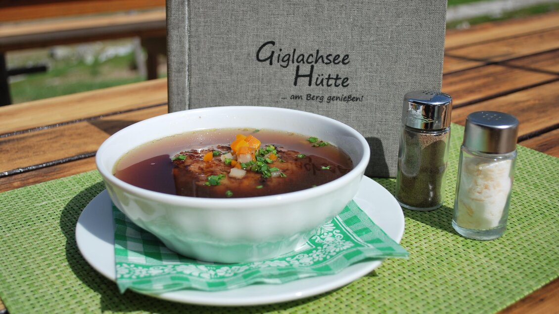 Eine Schüssel Suppe mit buntem Gemüse steht auf einem Tisch. Daneben sind Salz- und Pfefferstreuer sowie ein Menübuch platziert. | © Giglachseehütte