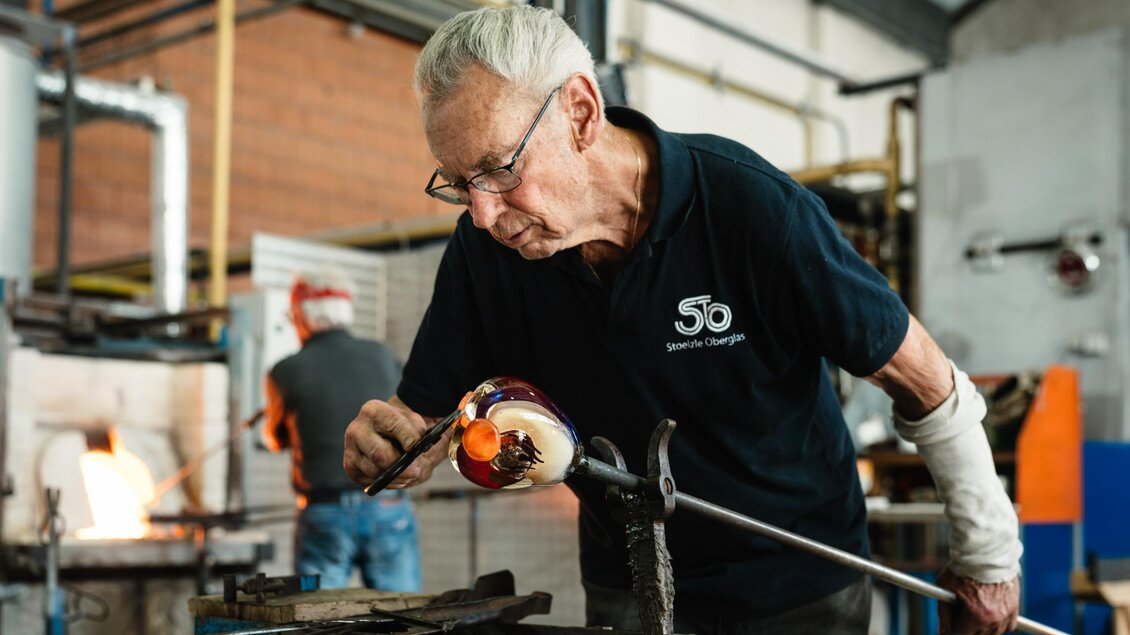 Ein Glasmacher formt mit einem Werkzeug ein farbiges Glasobjekt an der Glasmacherpfeife. | © Bernd Grosseck