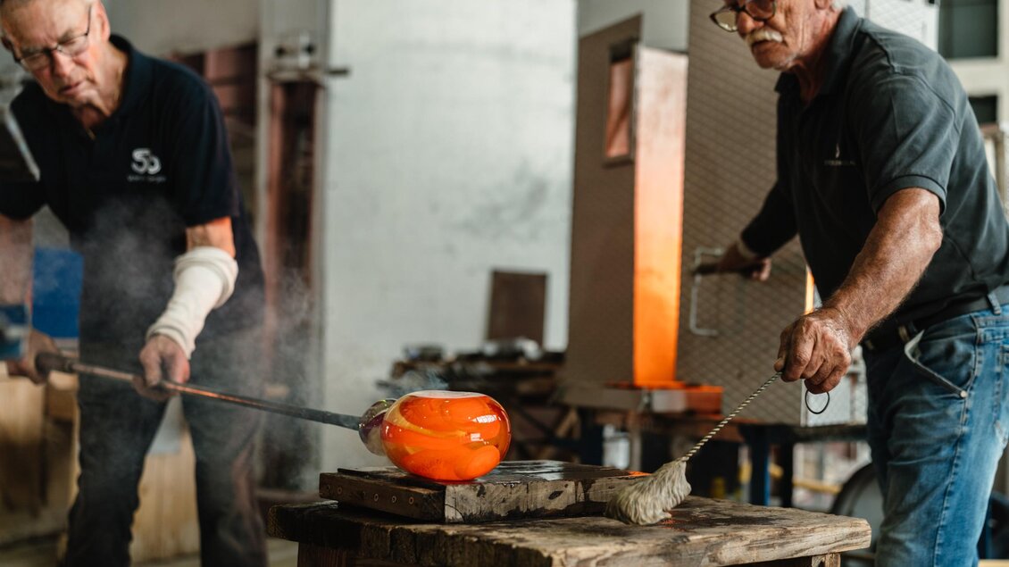 Zwei Glasmacher bearbeiten ein orange glühendes Glasobjekt in der Glashütte in Bärnbach. | © Bernd Grosseck