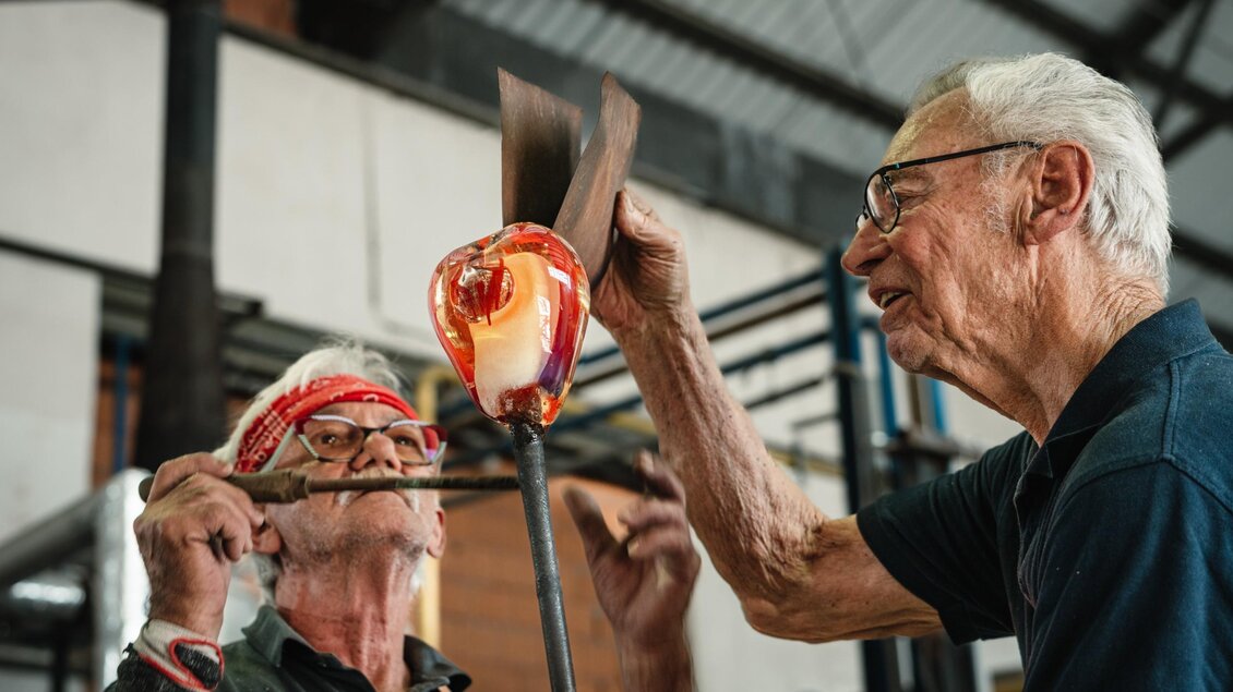 Zwei Glasmacher formen gemeinsam ein glühendes Glasobjekt mit verschiedenen Werkzeugen in Bärnbach. | © Bernd Grosseck