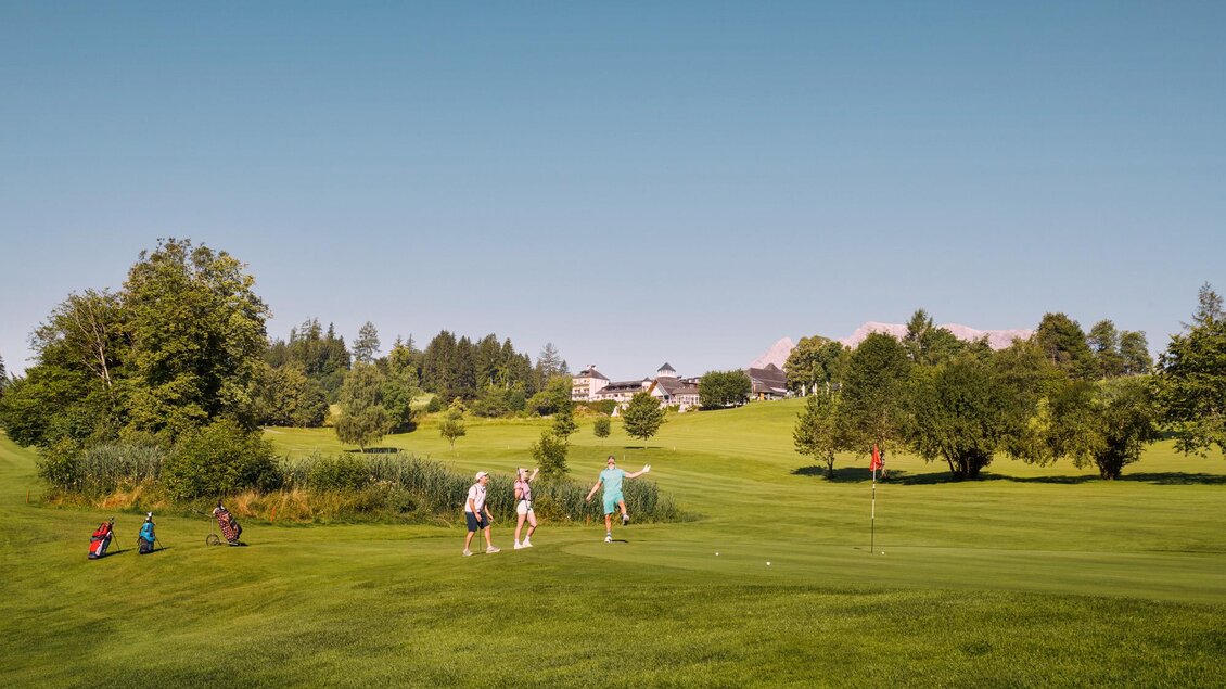 Eine grüne Golfanlage mit mehreren Spielern im Vordergrund. Im Hintergrund sind Bäume und Gebäude sichtbar unter einem klaren blauen Himmel. | © IMLAUER Hotel Schloss Pichlarn
