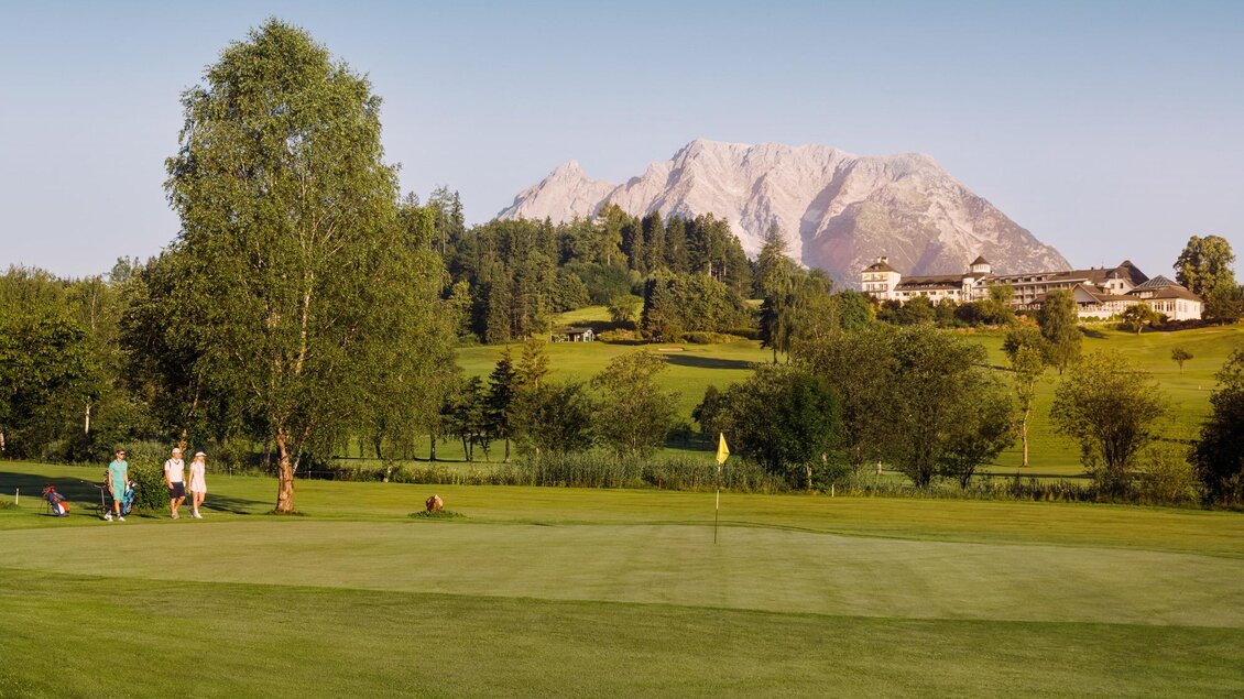 Eine wunderschöne Golfanlage mit dem Blick auf majestätische Berge im Hintergrund. Menschen spielen Golf auf einer grünen Wiese, umgeben von Bäumen und sanften Hügeln. | © Edy Gröger