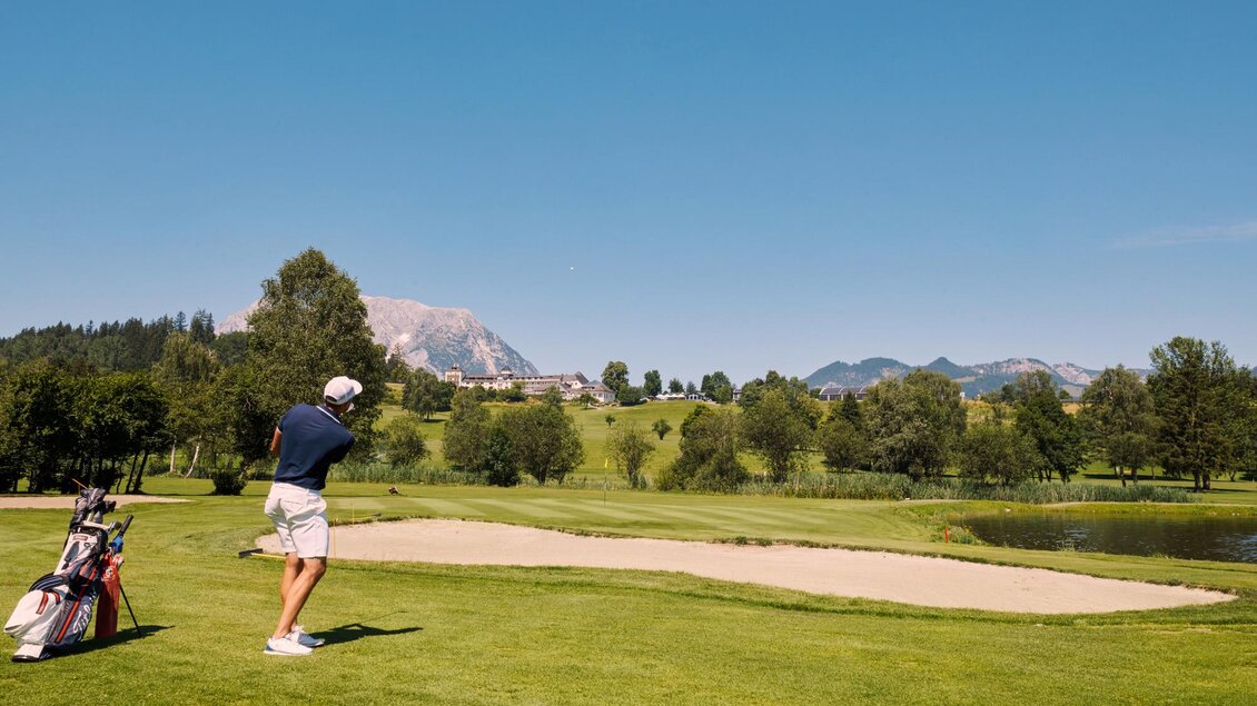 Ein Golfspieler schlägt einen Ball auf einem schönen Golfplatz mit Bergen im Hintergrund. Der Himmel ist klar und die Landschaft ist grün und gepflegt. | © Edy Gröger