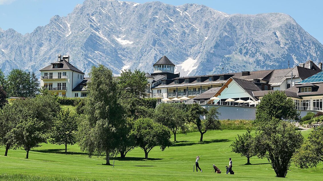 Eine malerische Landschaft mit einem Golfplatz im Vordergrund und majestätischen Bergen im Hintergrund. Im Bild erscheinen Bäume und einige Golfer. | © Golf & Country Club Schloss Pichlarn
