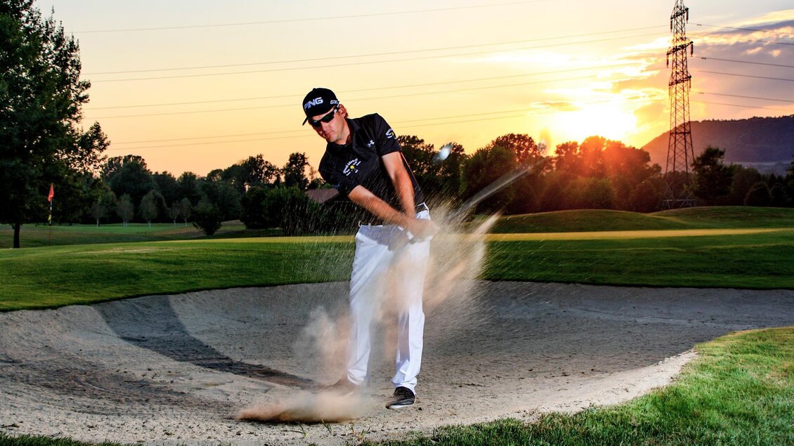 Ein Golfspieler schlägt aus einem Bunker, während die Sonne im Hintergrund untergeht. Die Landschaft ist grün mit Bäumen und einer beeindruckenden Abendstimmung. | © GV Murstätten