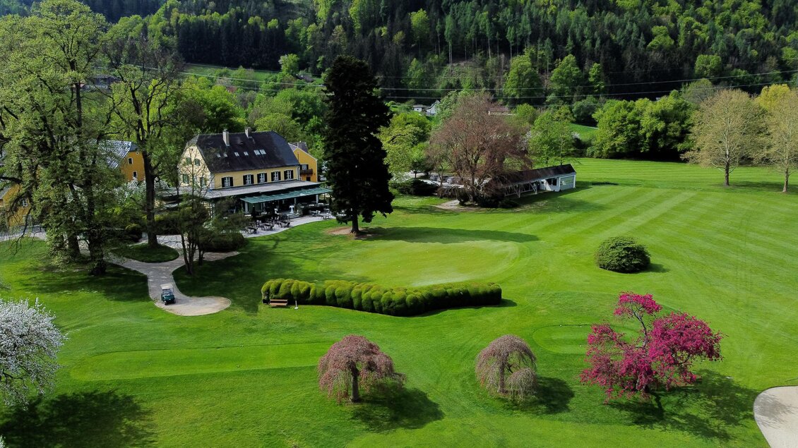 Luftaufnahme des Clubhauses und der gepflegten Golfanlage des Golfclubs Murhof mit blühenden Bäumen in Frohnleiten.  | © Golferlebnis Steiermark - Manuel Wiedner
