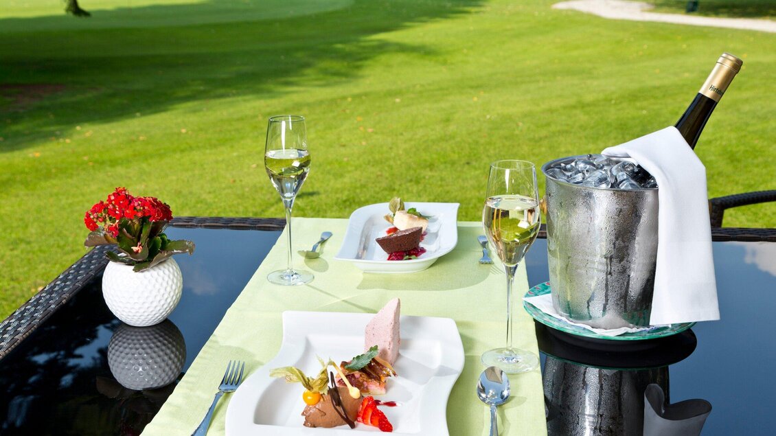 Ein elegantes Außentisch-Setup mit feinem Essen und Weingläsern. Im Hintergrund sieht man eine grüne Wiese und einen Kübel mit Eis für den Sekt. | © TV Region Graz - Rene Vidalli