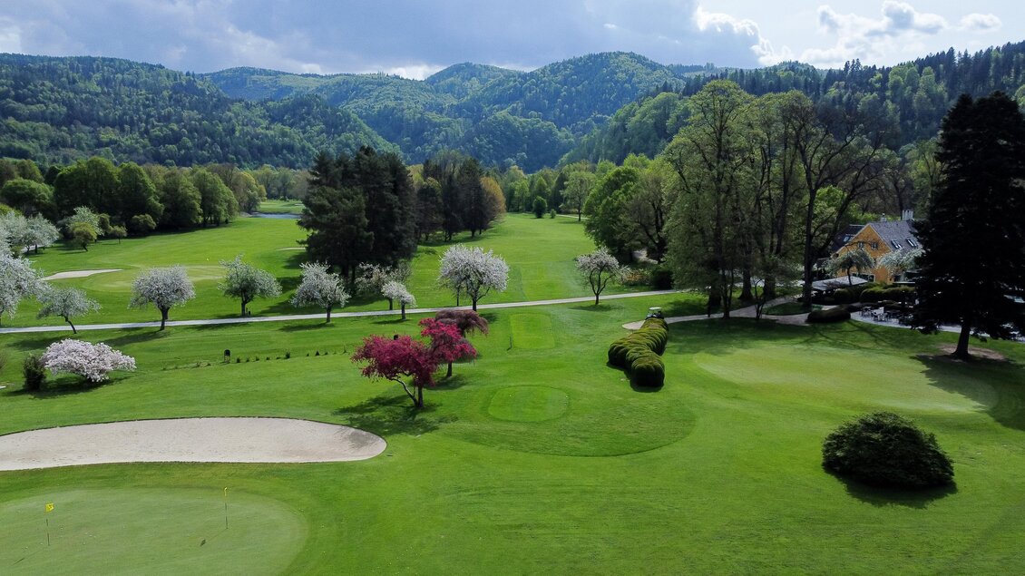 Blick über den Golfclub Murhof mit Fairways, blühenden Bäumen, Clubhaus und bewaldeten Hügeln im Hintergrund in Frohnleiten. | © Golferlebnis Steiermark - Manuel Wiedner