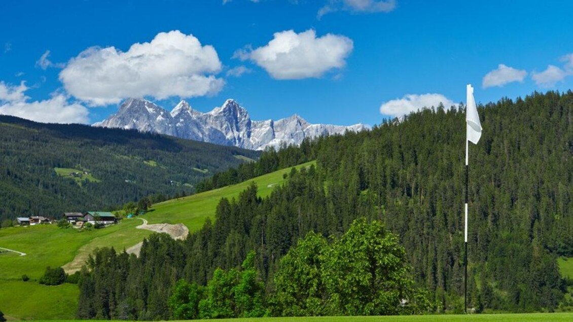 Eine malerische Landschaft mit hohen Bergen und grünen Wäldern. Im Vordergrund steht eine Fahne auf einem Golfplatz. | © Golfclub Radstadt
