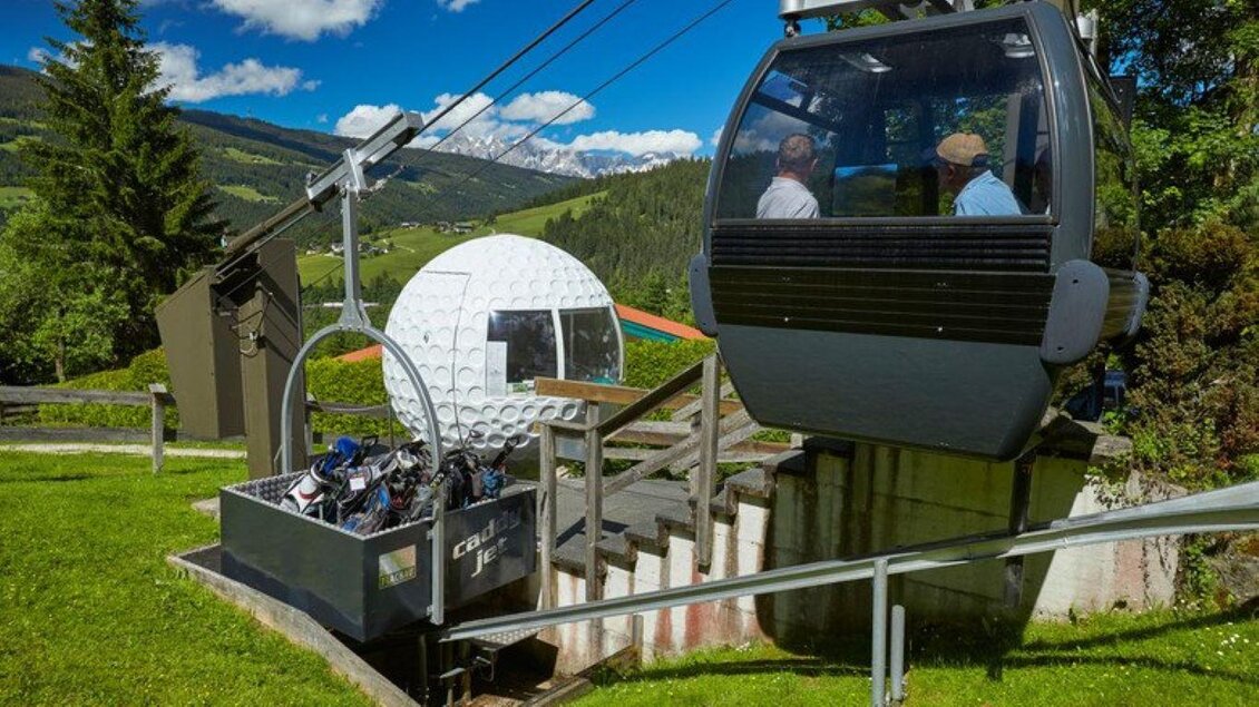 Eine Seilbahn mit Passagieren fährt über eine grüne Wiese. Im Vordergrund ist ein geschlossener Kabinenwagen zu sehen, der mit Geräten gefüllt ist. | © Golfclub Radstadt