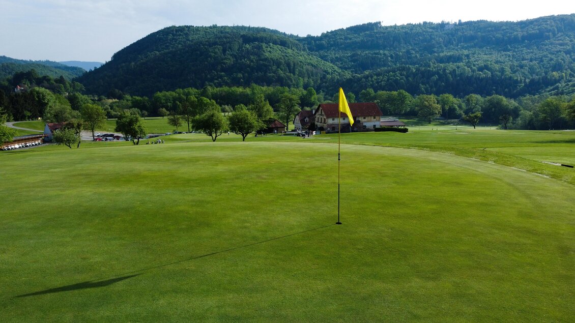 Blick über das Green mit Fahne zum Clubhaus des Golfclubs Thalersee vor bewaldeten Hügeln in Thal. | © Golferlebnis Steiermark - Manuel Wiedner