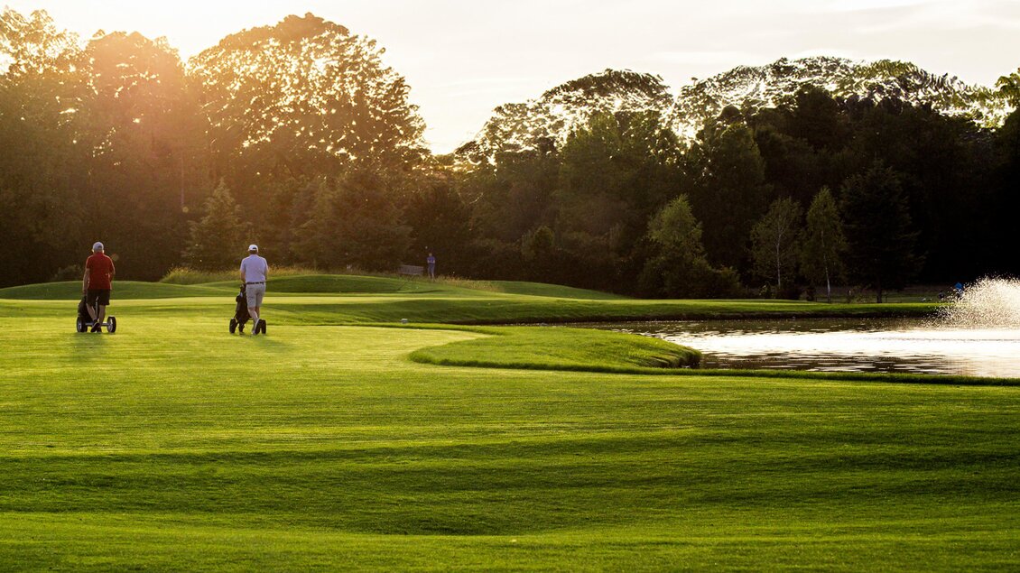 Zwei Golfer mit Trolleys auf dem Fairway neben einem Wasserhindernis im Golfclub Grazer MurAuen bei Abendsonne in Gössendorf.  | © Golferlebnis Steiermark - Manuel Wiedner