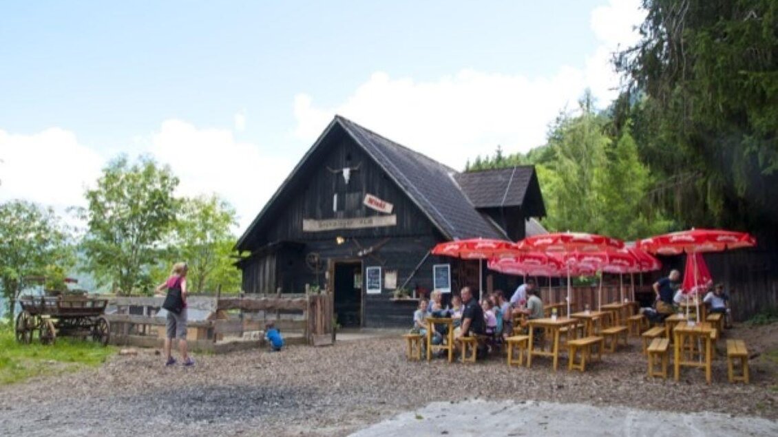 Ein gemütliches Restaurant in den Bergen mit Holzgebäude und stilvollen Terrassen. Es gibt mehrere Tische unter roten Sonnenschirmen, die Gäste genießen die Atmosphäre. | © Gröbminger Alm