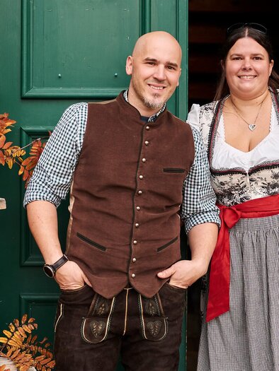Ein Paar in traditioneller bayerischer Kleidung steht vor einer grünen Tür. Im Hintergrund sind herbstliche Dekorationen zu sehen.