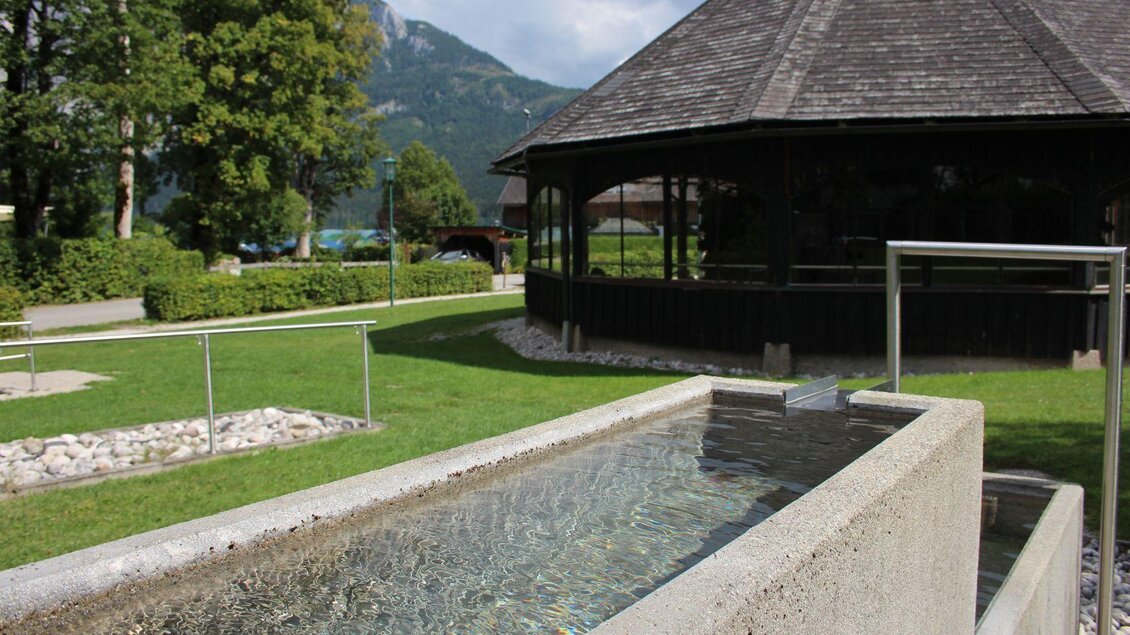 Ein modernes Wasserbecken aus Beton in einer grünen Umgebung. Im Hintergrund ist ein Holzpavillon und Berge sichtbar. | © Viola Lechner