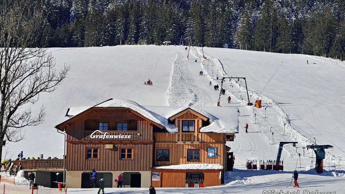 <p>Die Skihütte Grafenwiese neben dem Grafenwiesenlift in der schneebedeckten Winterlandschaft. Es sind einige Skifahrer am Schlepplift zu sehen.</p> | © Melanie Fritz_Steirisch Sche