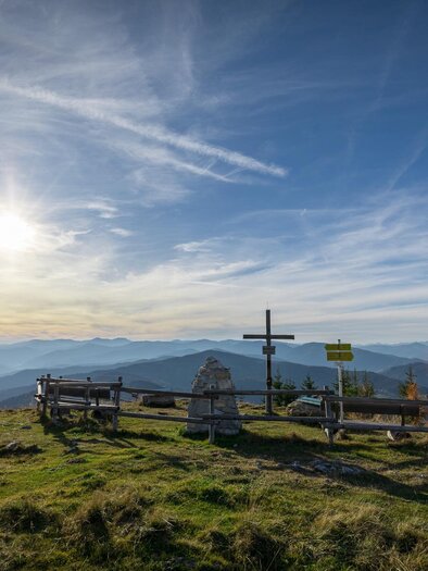 Eine schöne Berglandschaft mit einem Holzgeländer und einem Kreuz. Der Himmel ist klar mit einigen Wolken, während die Sonne aufgeht. | © Tourismusverband Murau