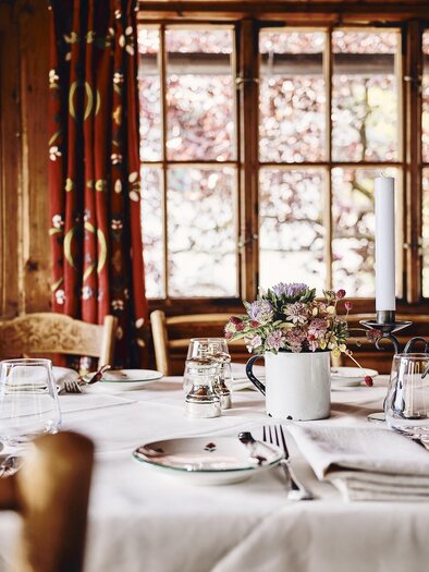 A cozy table with a white tablecloth and dishes in a wooden room. Flowers in a cup and large windows create a warm atmosphere. | © Armin Walcher
