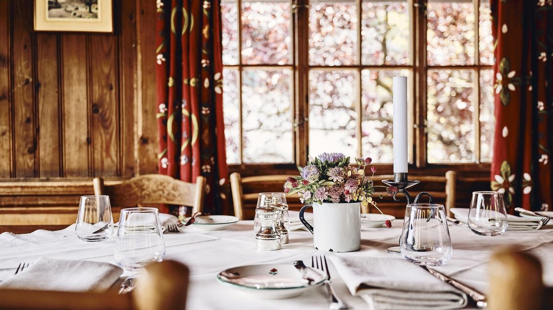 Ein gemütlicher Tisch mit weißer Tischdecke und Geschirr in einem Holzraum. Blumen in einer Tasse und große Fenster sorgen für eine warme Atmosphäre. | © Armin Walcher