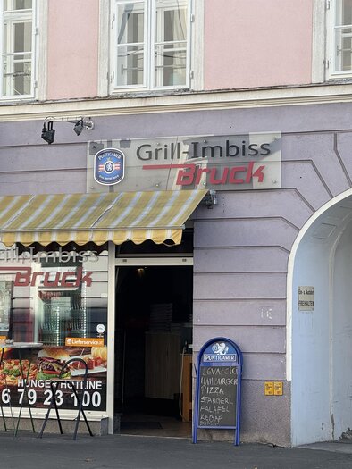 A grill snack bar with a yellow sun umbrella and an inviting facade. The signs display the name "Grill-Imbiss Dück" and the contact number. | © TV Hochsteiermark