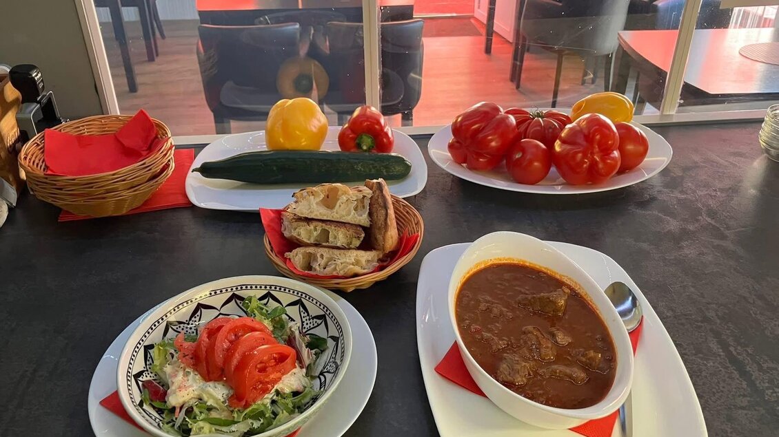Ein Tisch mit einer Auswahl an frischen Lebensmitteln. Auf dem Teller befinden sich eine Suppe, ein Salat mit Tomaten und Brot sowie einige frische Tomaten, Gurken und Obst.