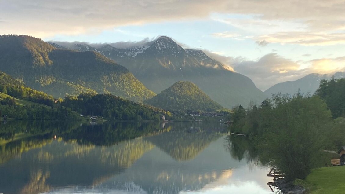 Der ruhige Grundlsee, aufgenommen vom Surfplatz am Gaiswinkel. Umgeben von Bergen des Ausseerland Salzkammergutes und sommerlichem Grün spiegeln sich die Wolken sanft im Wasser und schaffen eine friedliche Atmosphäre. | © Bettina Scheck