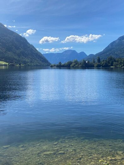 The tranquil lake Grundlsee photographed from the direction of Gößl, surrounded by the mountains of the Ausseerland Salzkammergut region. The sky is clear and blue, and the water reflects the beautiful landscape. | © Bettina Scheck