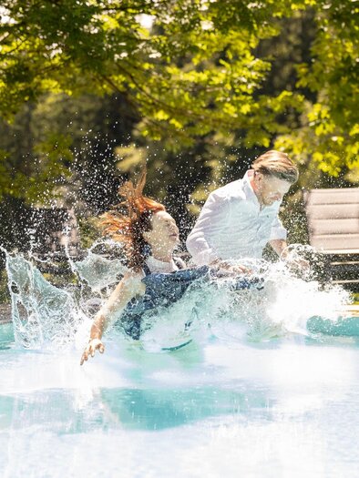 Ein Mann und eine Frau springen lachend in einen Pool und spritzen Wasser. Die Umgebung ist grün und sonnig, was eine fröhliche Atmosphäre schafft. | © Gut Moser