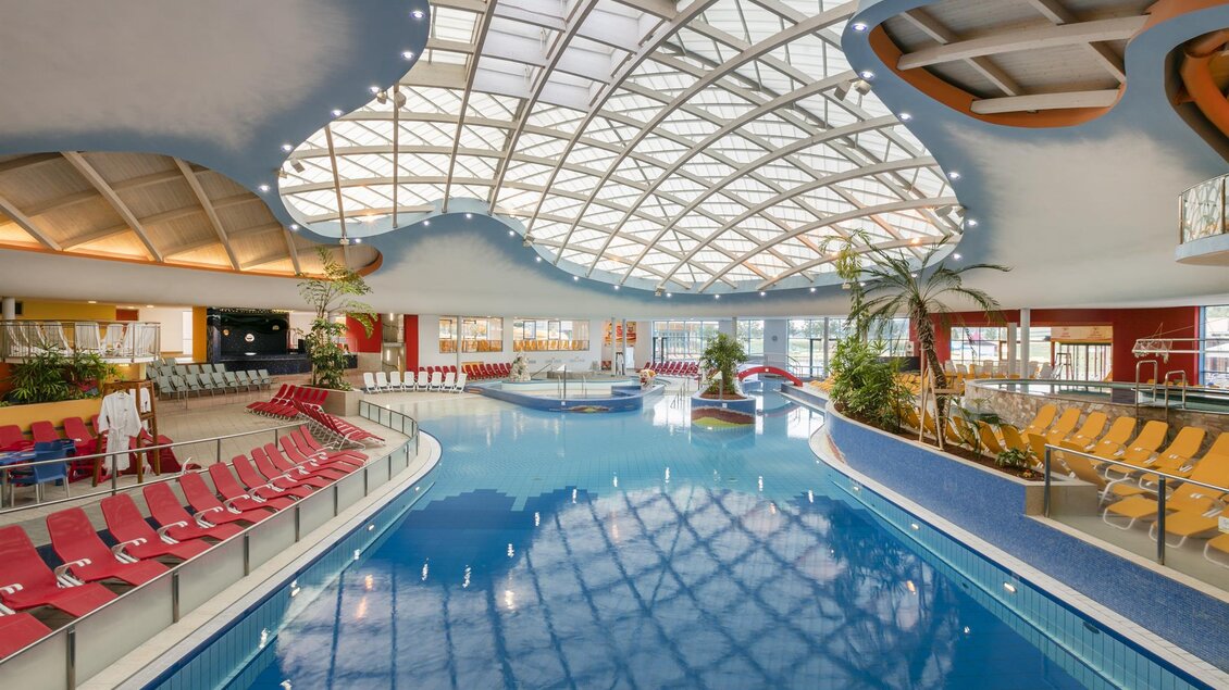Ein modernes Hallenbad mit einem großen blauen Wasserbecken und einem gewölbten Glasdach. Die Umgebung ist mit Liegen und Pflanzen dekoriert. | © H2O-Hotelterme GmbH