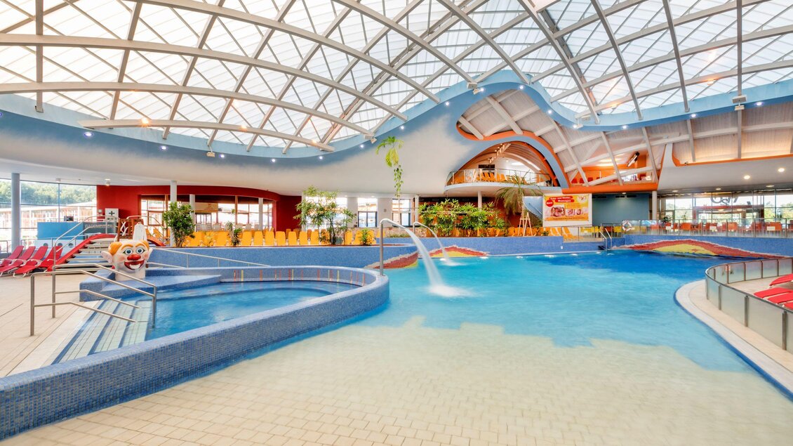 Ein modernes Hallenbad mit gläsernem Dach und blauen Wasser. Es gibt Rutschen und Liegen für Besucher. | © H2O-Hoteltherme GmbH