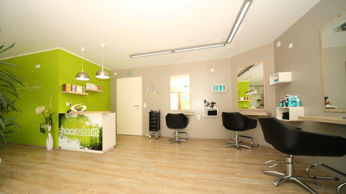 Ein moderner Friseursalon mit hellen Wänden und stylischen Stühlen. Die Einrichtung ist schlicht und freundlich, mit einer grünen Akzentwand. | © Haarschnitt