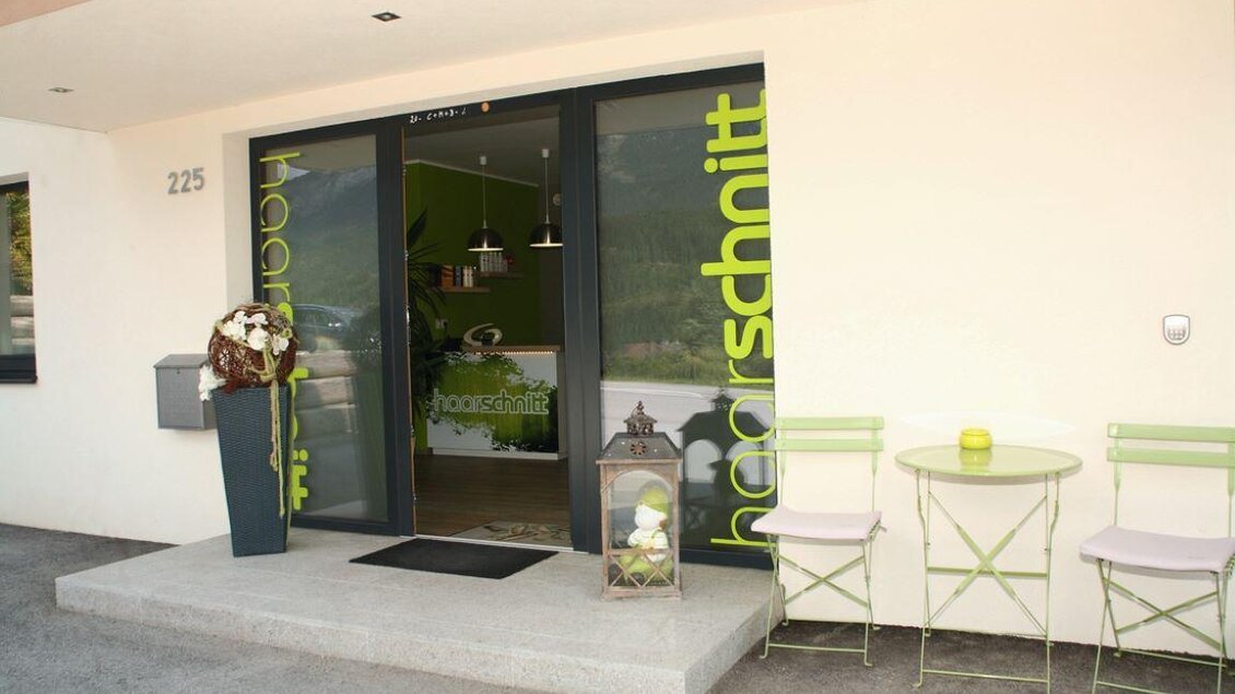 Eingang zu einem modernen Friseursalon mit großen Fenstern. Draußen stehen ein Tisch und Stühle bereit. | © Haarschnitt