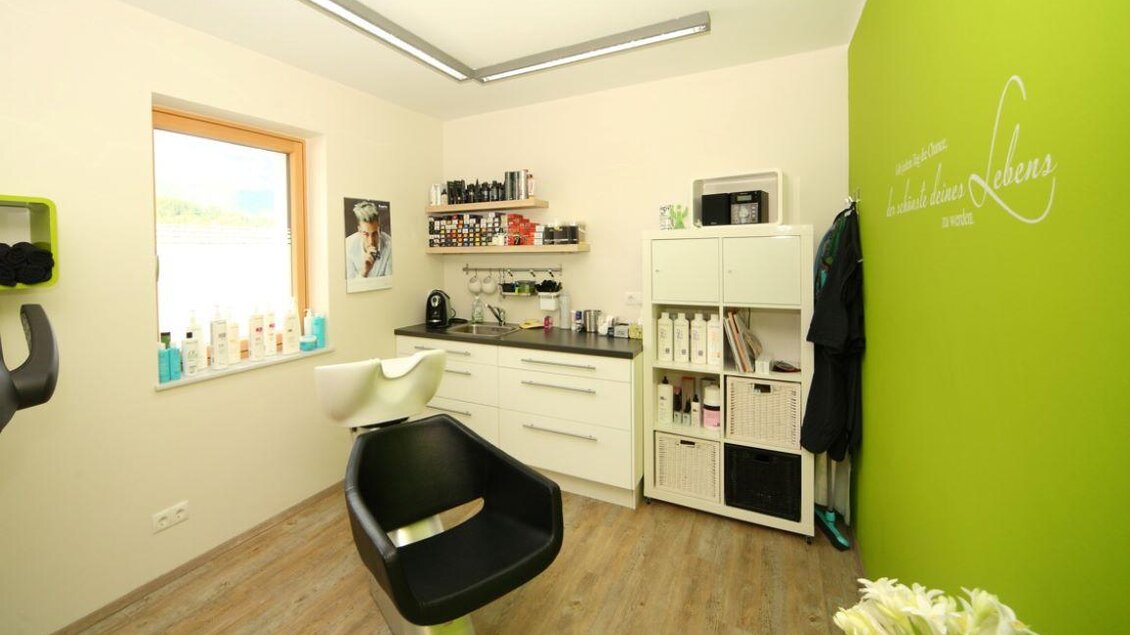 Ein helles Friseurstudio mit einem modernen Friseurstuhl und einem gut organisierten Arbeitsplatz. An der Wand befindet sich eine grüne Akzentfarbe, und Regale sind mit Pflegeprodukten gefüllt. | © Haarschnitt