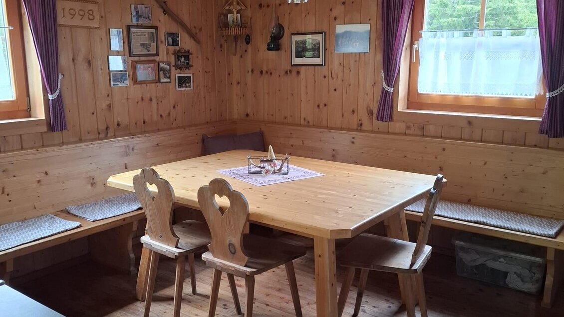 Ein rustikaler Essbereich mit einem großen Holztisch und Stühlen. Die Wände sind aus Holz, und die Fenster sind mit Vorhängen dekoriert. | © Rinnhofer
