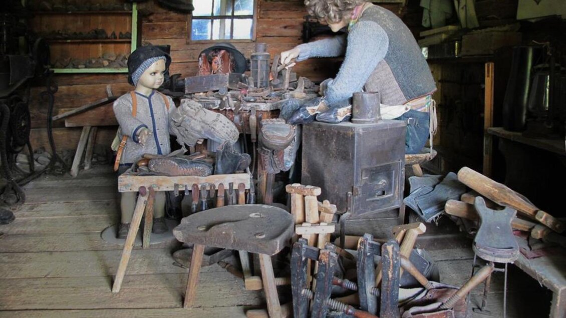 Ein Werkraum mit einem Handwerker, der an einem Tisch arbeitet, umgeben von alten Werkzeugen. Neben ihm sitzt eine Holzfigur eines Kindes. | © Gemeinde St. Veit in der Südsteiermark