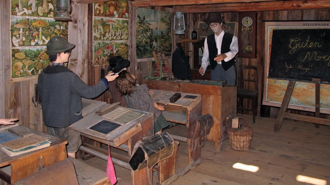 Ein historisches Klassenzimmer mit Holzmöbeln und alten Tafeln. Schüler und ein Lehrer sind in einer Lernumgebung aus der Vergangenheit dargestellt. | © Gemeinde St. Veit in der Südsteiermark