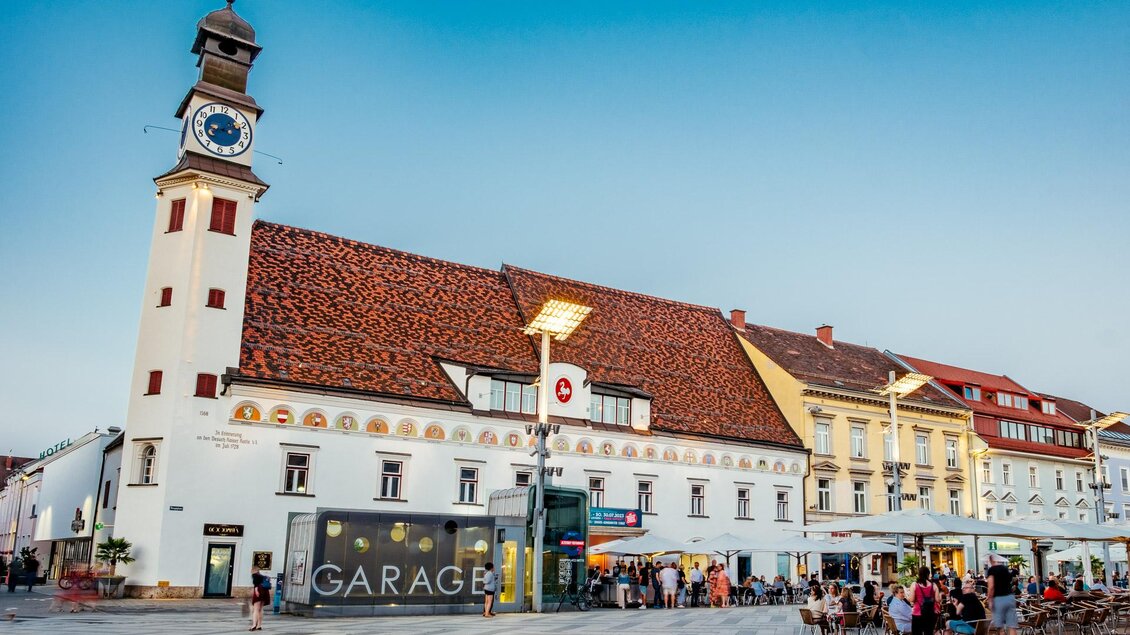 Ein historisches Gebäude mit einer Uhrturm und bunten Wandmalereien. Vor dem Gebäude sitzen Tische und Stühle im Freien. | © TVB Erzberg-Leoben