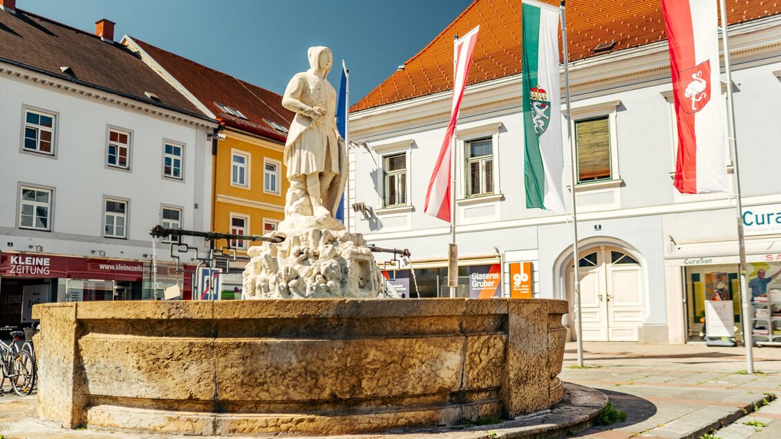 Ein Paar entspannt an einem Brunnen in einer lebhaften Stadt. Bunte Gebäude und eine angenehme Atmosphäre umgeben sie. | © TVB Erzberg-Leoben