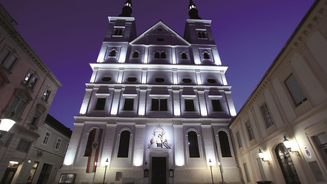 Eine malerische Straße mit Geschäften und einer beeindruckenden Kirche im Hintergrund. Der Himmel ist bewölkt und die Gebäude sind nachtbeleuchtet. | © TVB Erzberg-Leoben