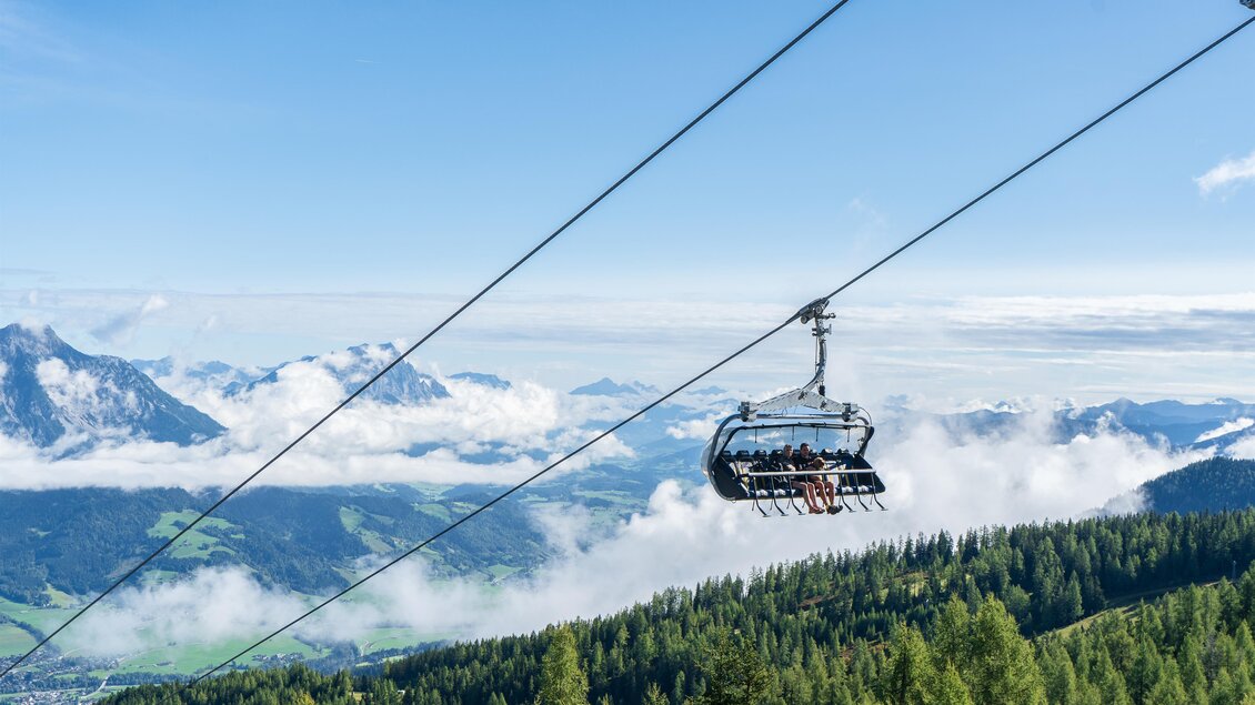 Eine Seilbahn schwebt über grüne Wälder und Wolken. Im Hintergrund sind majestätische Berge und ein blauer Himmel zu sehen.