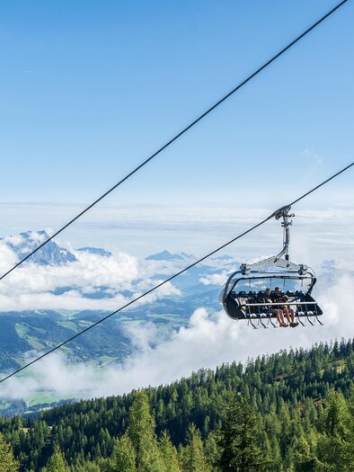 Eine Seilbahn schwebt über grüne Wälder und Wolken. Im Hintergrund sind majestätische Berge und ein blauer Himmel zu sehen.