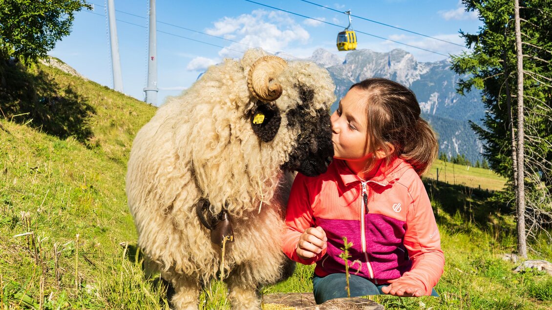 Ein Mädchen küsst ein Schaf auf einer grünen Wiese in den Bergen. Im Hintergrund sind Seilbahnen und eine schöne Berglandschaft zu sehen.
