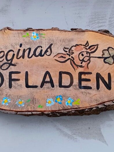Ein dekoratives Holzschild mit dem Schriftzug „Reginas Hofladen“. Das Schild zeigt auch eine Zeichnung einer Kuh und bunte Blumen. | © Regina Pehringer