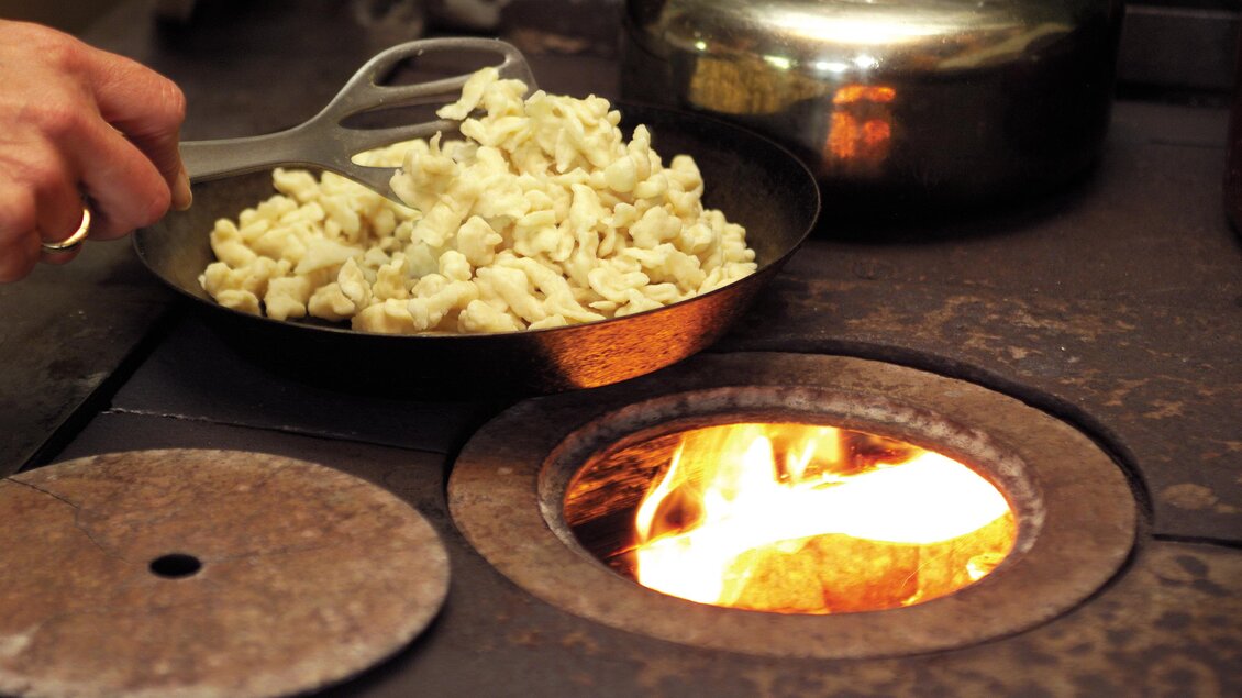 Eine Hand hält einen Spatel über einer Pfanne mit gekochtem Teig. Darunter brennt ein offenes Feuer in einem Herd. | © Hotel DER HECHL