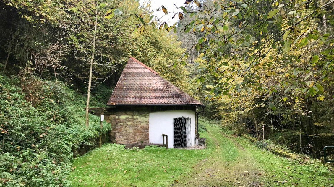 Ein kleiner, runder Bau mit rotem Dach steht am Rand eines Waldwegs. Die Umgebung ist von grünen Bäumen und herbstlichen Farben geprägt. | © TV Südsteiermark