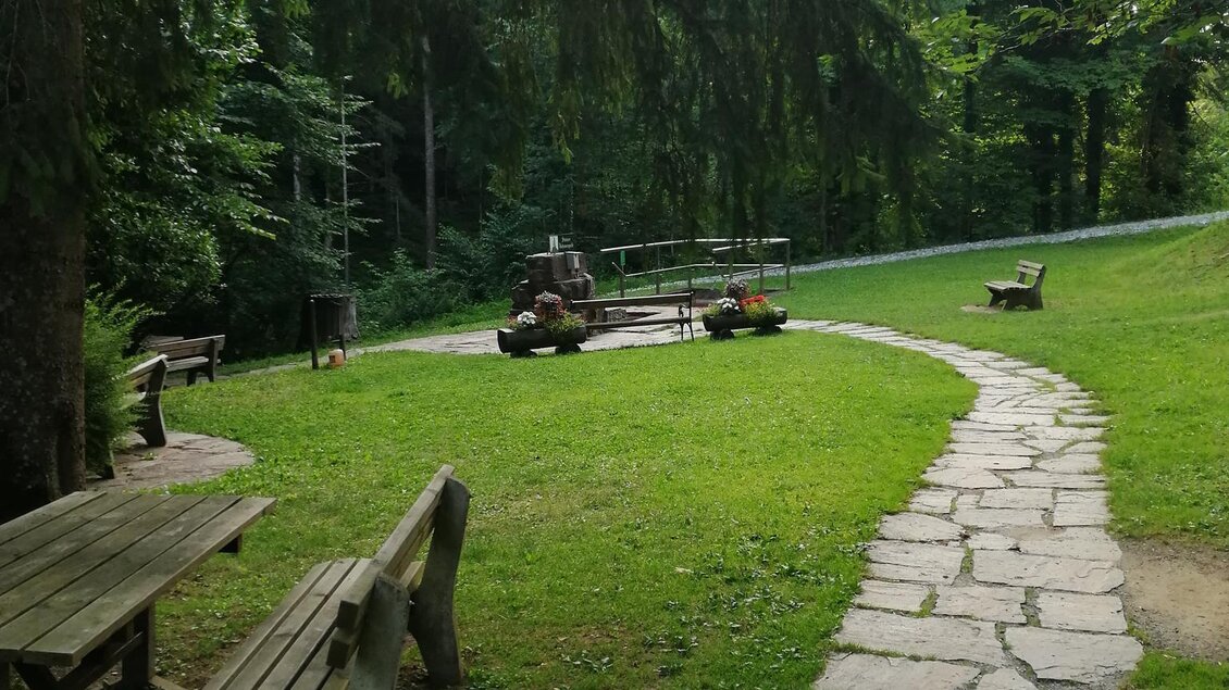 Ein gepflegter Garten mit einem gepflasterten Weg und einer Holzbank. Im Hintergrund sind Bäume und ein kleiner Rasenplatz zu sehen. | © TV Südsteiermark
