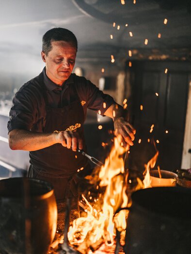 Ein Koch steht vor einem großen Feuer und rührt darin. Funken fliegen auf, während er die Speisen zubereitet. | © Oststeiermark Tourismus