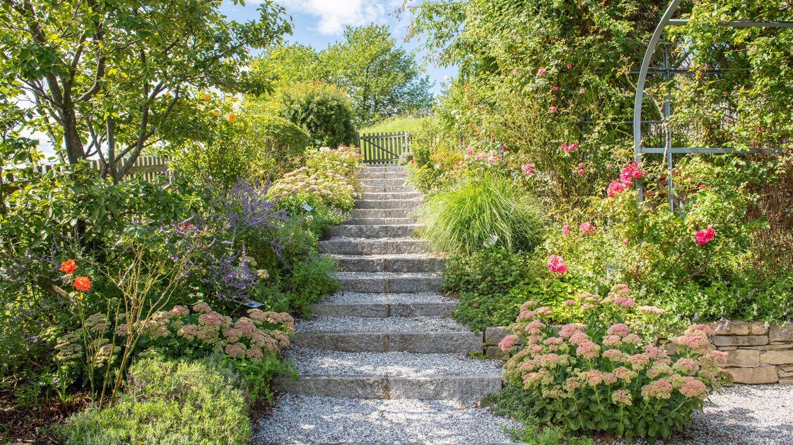 Ein schöner Garten mit blühenden Pflanzen und einer steinernen Treppe. Im Hintergrund ist ein Tor und grüne Bäume zu sehen. | © Helmut Schweighofer